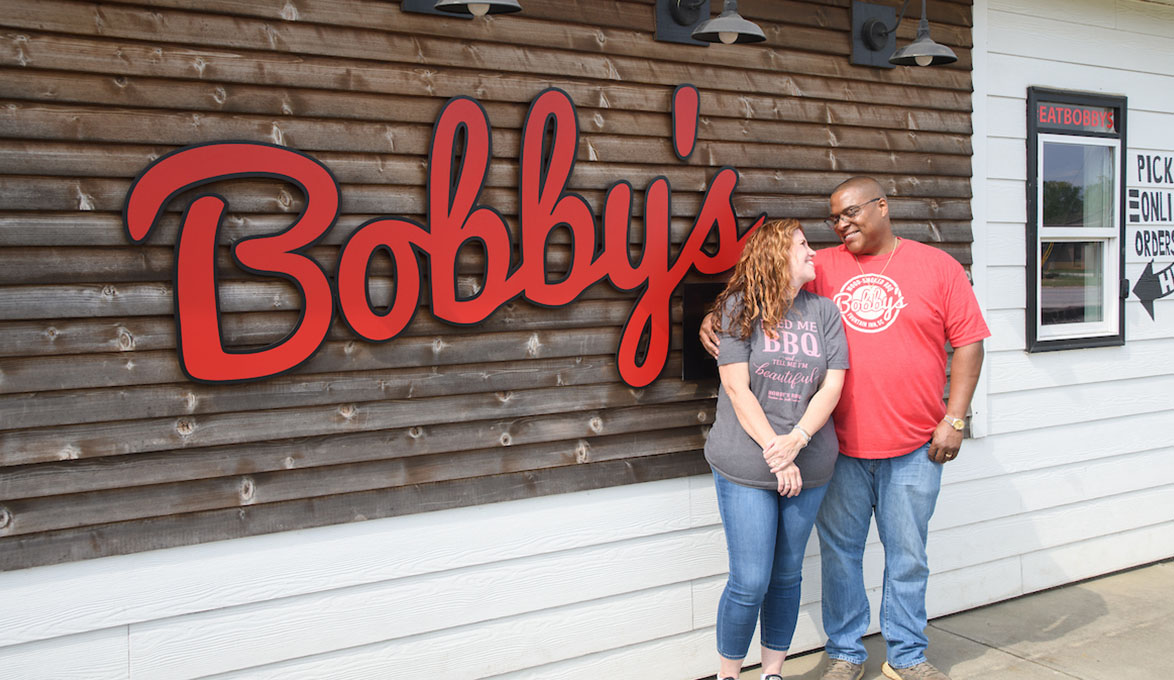Two People in front of Bobby's BBQ sign.